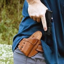 Close-up of a person lifting a blue shirt while drawing a black handgun from a brown leather OWB holster worn on the hip.